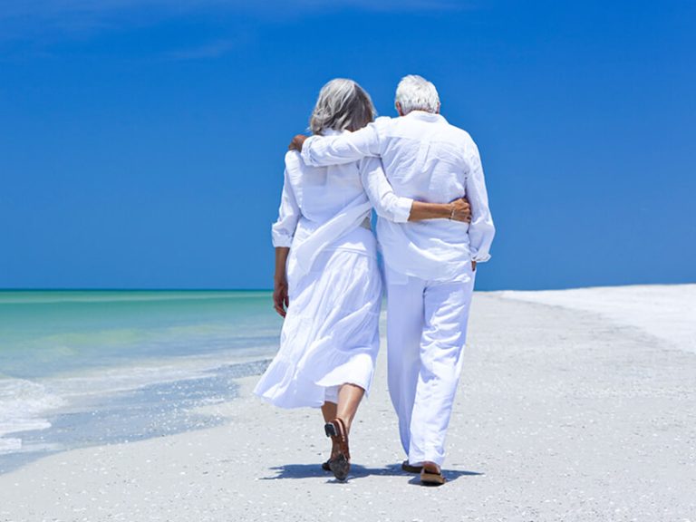 ηλικιωμένο ζευγάρι με λευκά ρούχα κάνει βόλτα στην παραλία